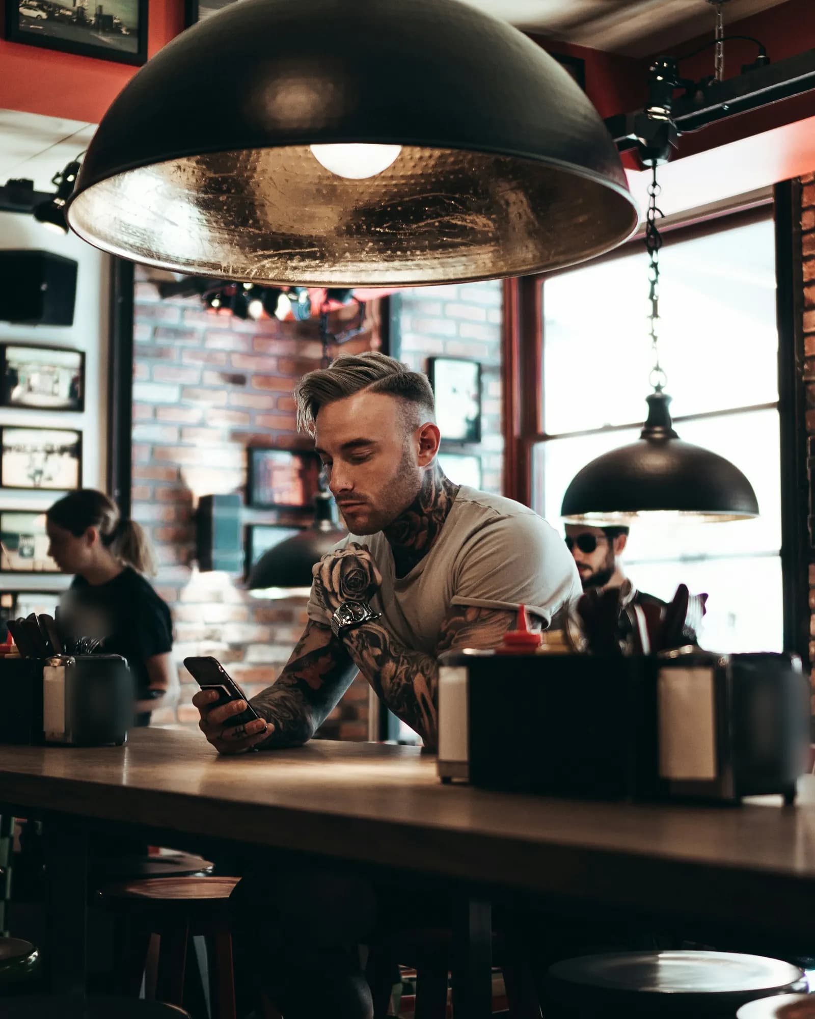 Un homme aux bras tatoués, accoudé au comptoir d’un bar aux murs de brique, lit son téléphone sous une grande suspension noire ; une serveuse passe derrière lui.