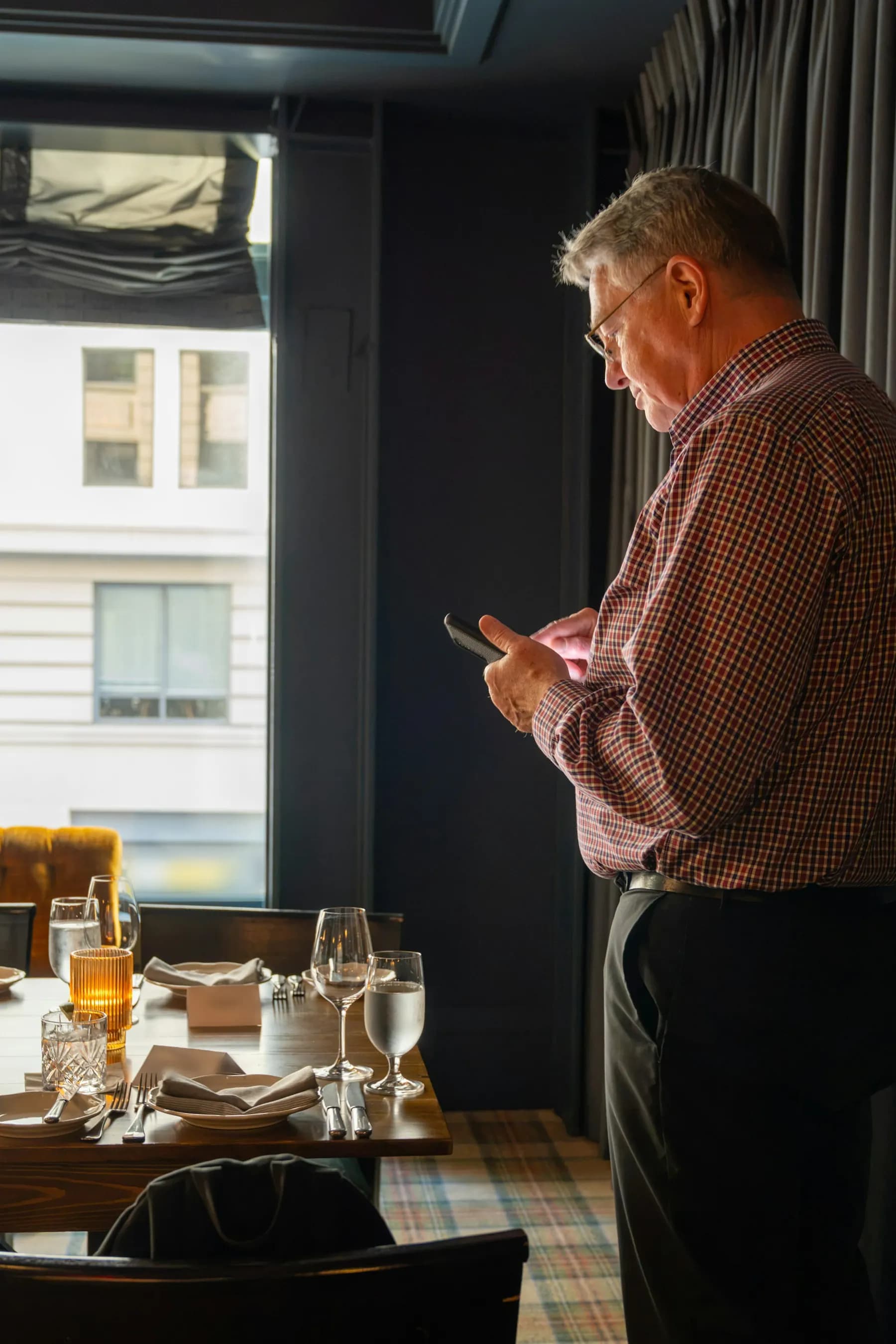 Dans une salle de restaurant encore vide, un homme en chemise à carreaux écrit sur son téléphone, près d’une table dressée.