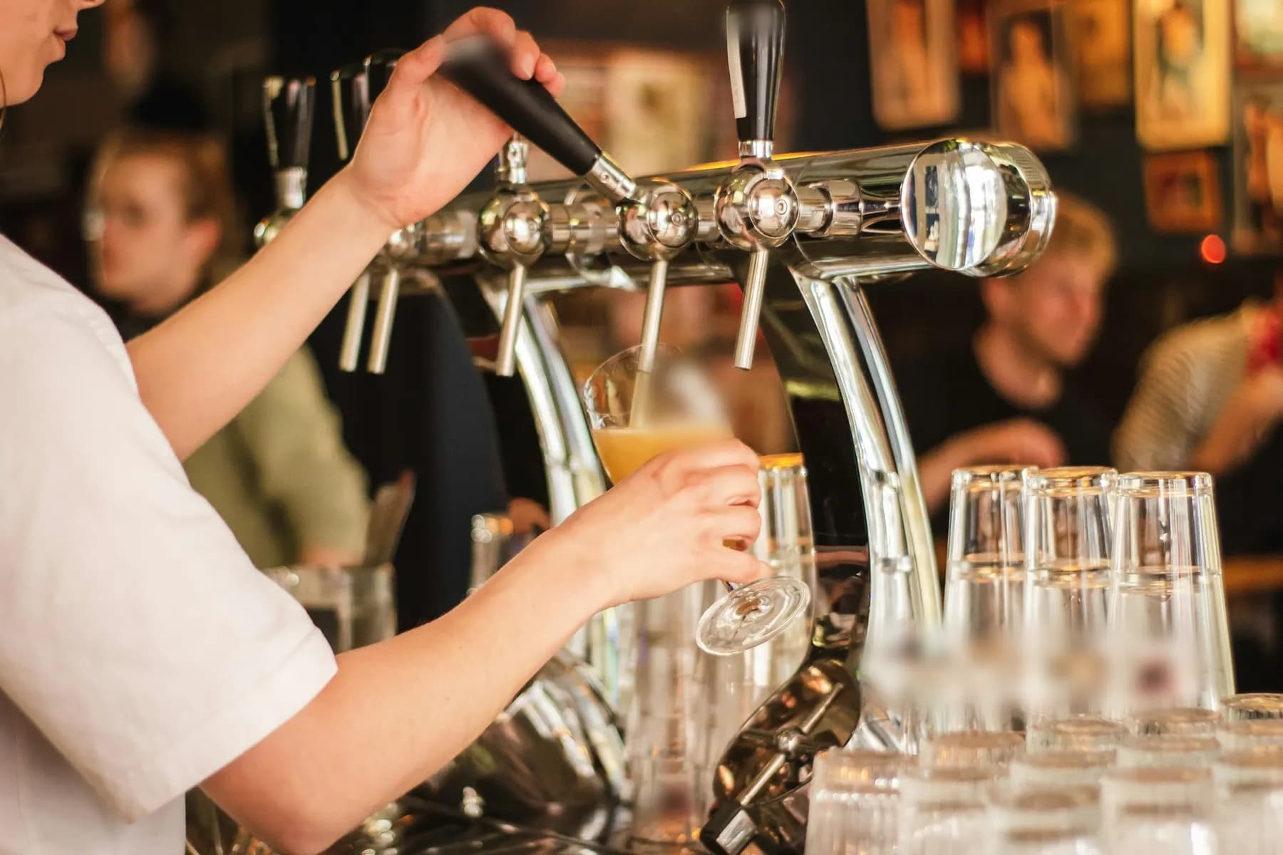 Au comptoir d’un bar, une serveuse tire une pression, devant une rangée de verres et des clients attablés.