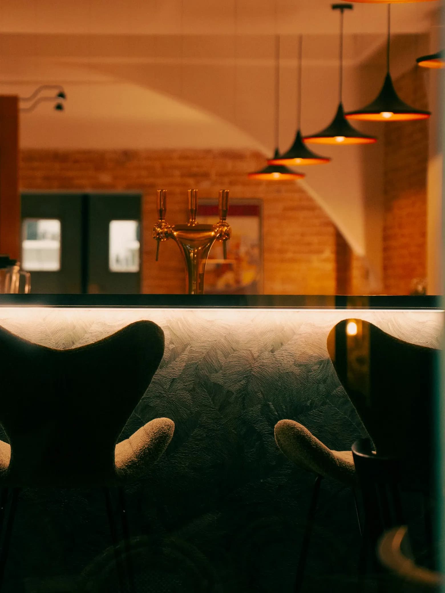 Un comptoir de bar vide avant l’ouverture, ses tabourets et sa tireuse sous des suspensions allumées.