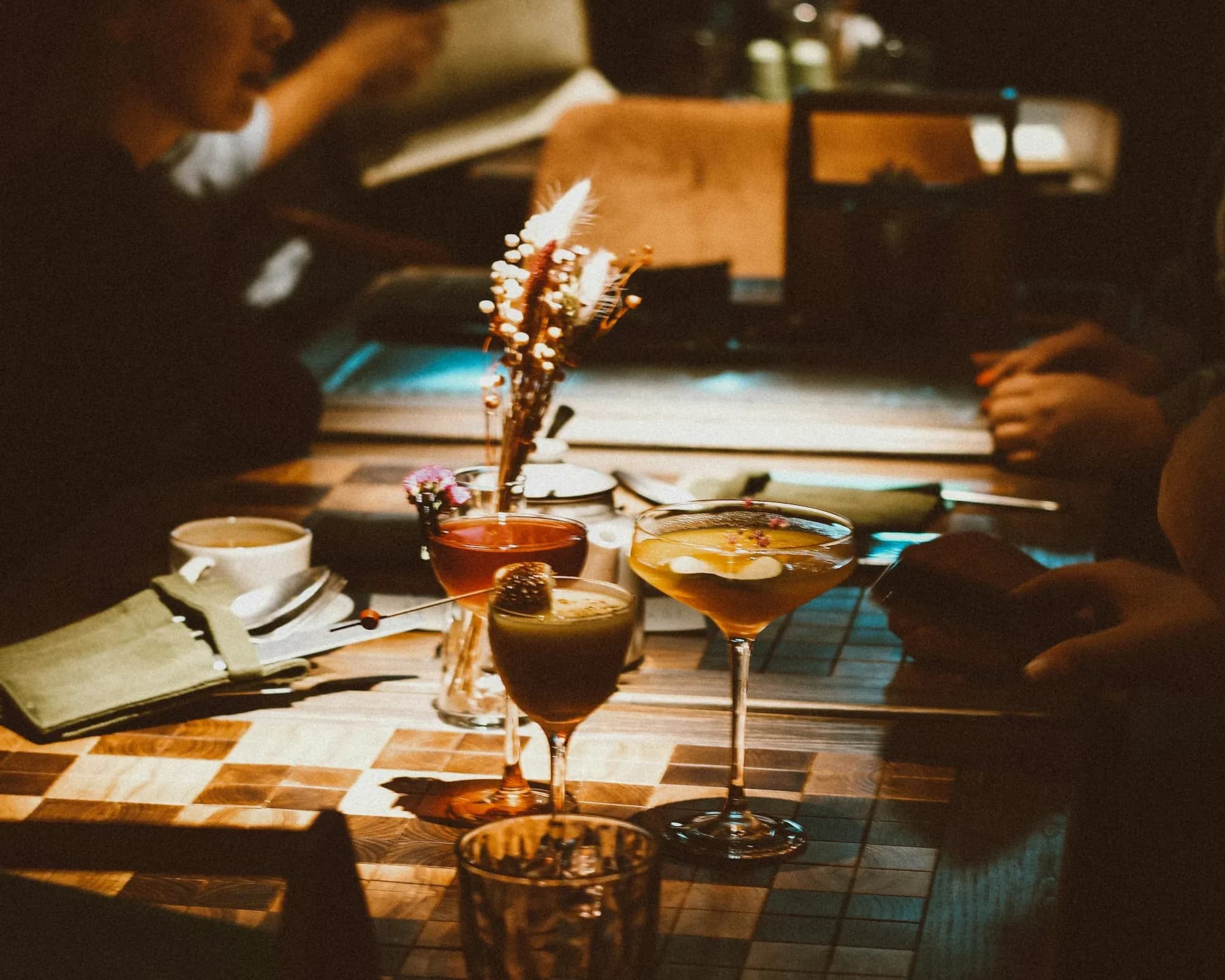 Trois cocktails sur une table en damier, dans la lumière chaude d’un bar, des clients attablés autour.