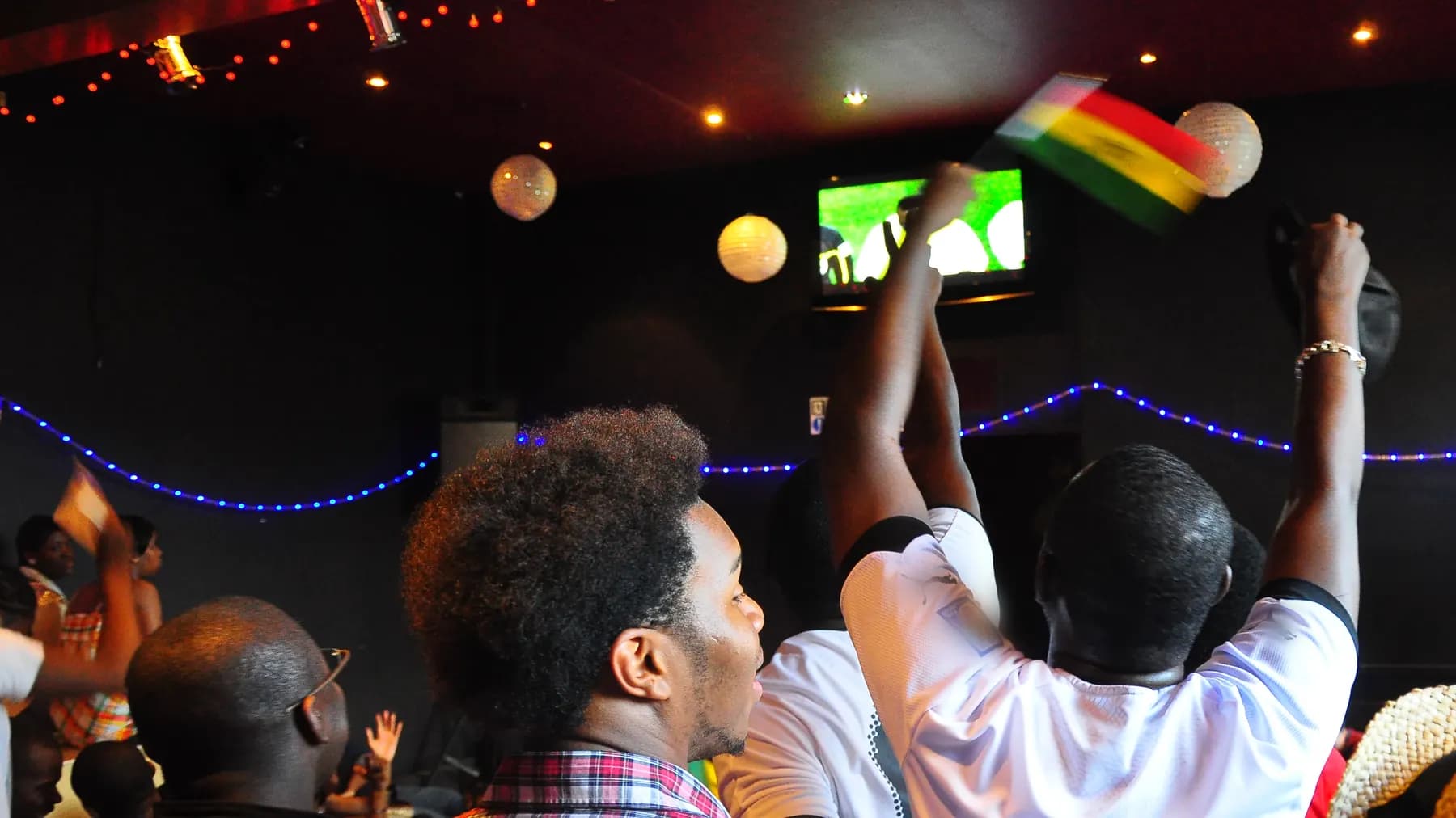 Dans un bar sombre aux lampions et aux guirlandes lumineuses, des supporters debout, de dos, lèvent les bras et agitent un drapeau devant l’écran où passe le match.