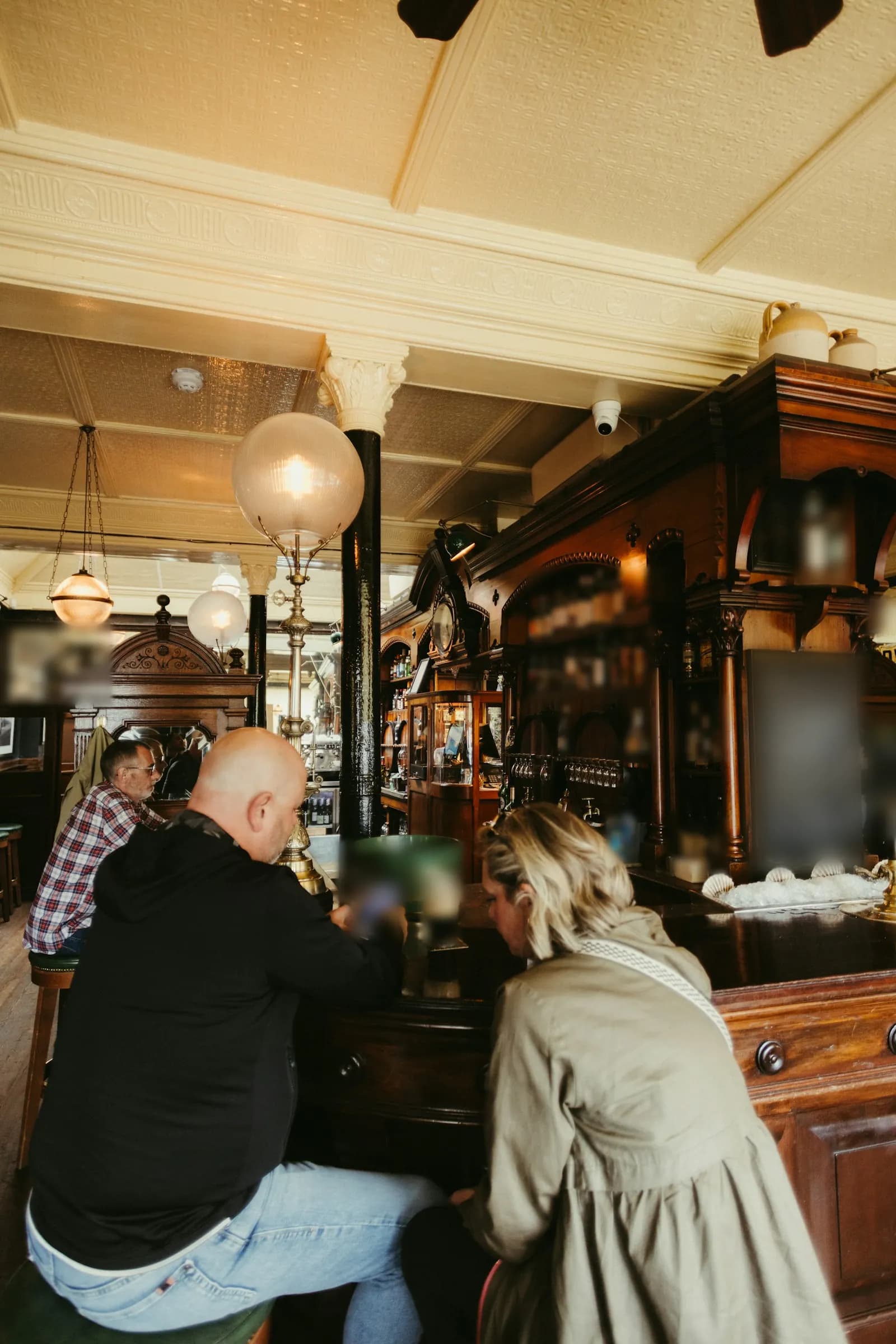 Dans un pub aux boiseries sombres, un homme assis au comptoir regarde son téléphone devant une pinte, une femme penchée à côté de lui, d’autres clients au fond.