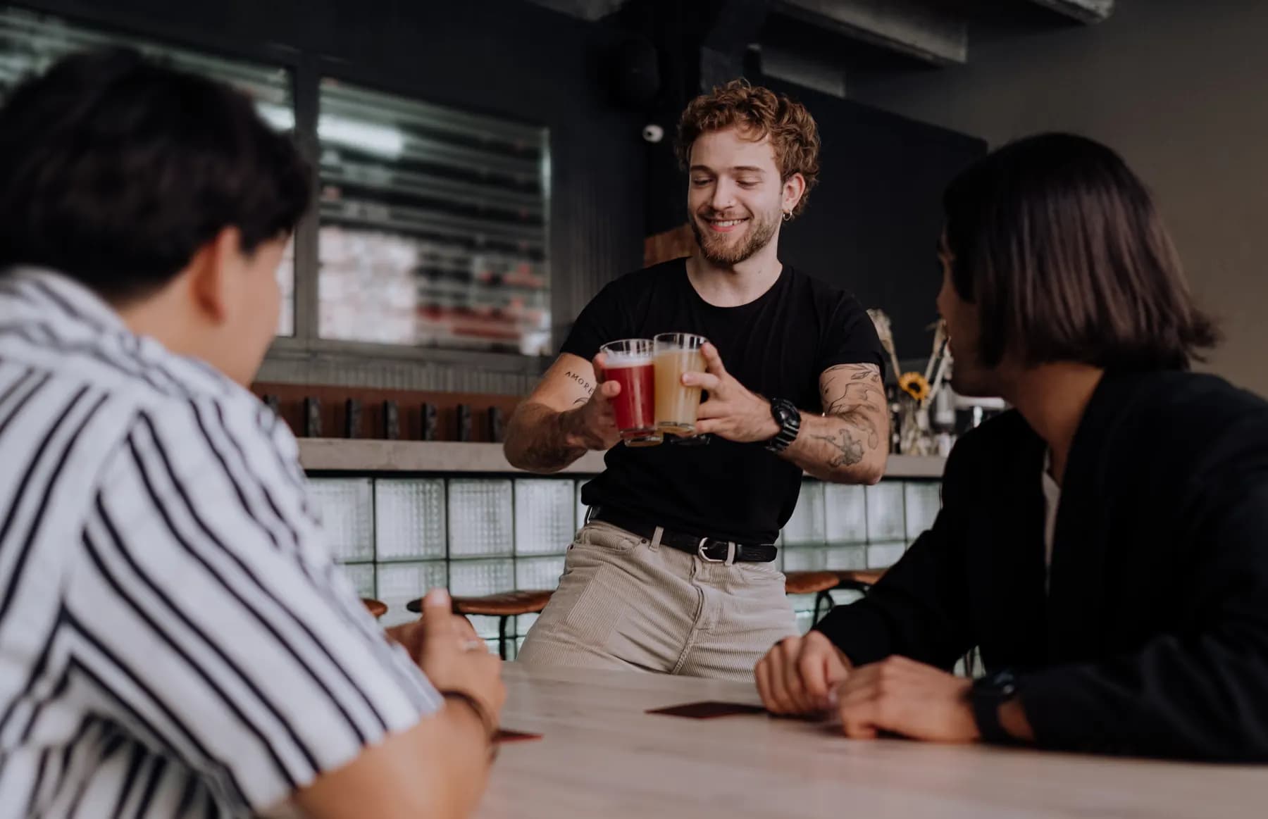 Dans un bar, devant le comptoir, un homme souriant en t-shirt noir tend deux bières, une rouge et une blonde, à deux clients assis à une table.