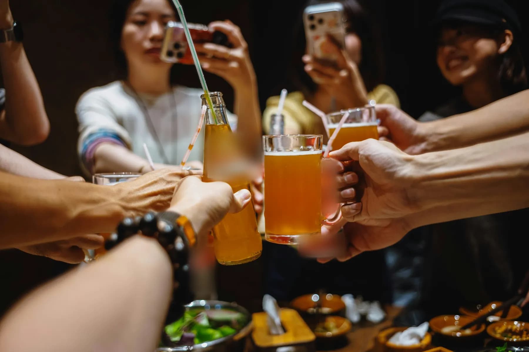 Autour d’une table, des amis trinquent, bouteille et chopes levées. Au fond, deux d’entre eux tiennent leur téléphone.