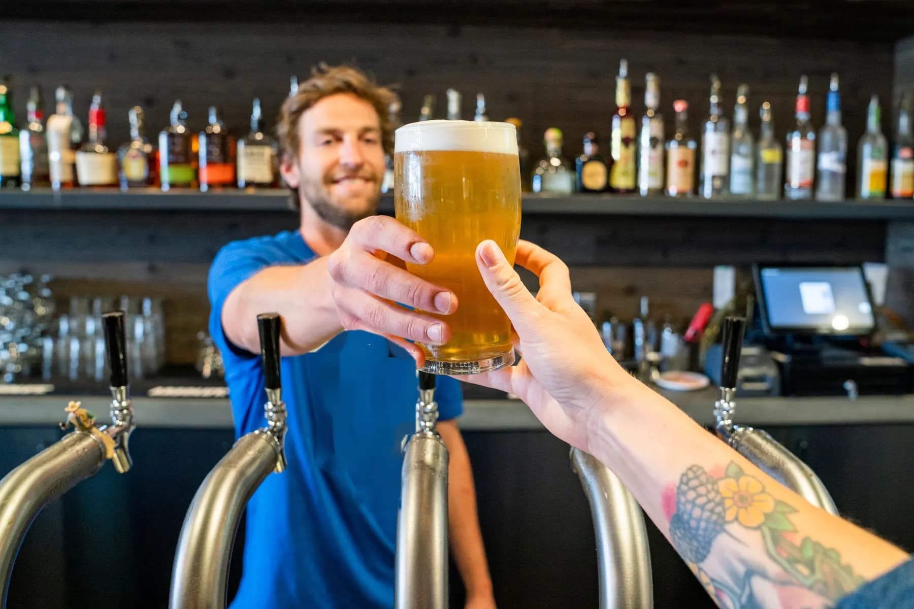 Au comptoir d’un bar, un barman tend une pinte de bière par-dessus les tireuses à la main d’un client, devant une rangée de bouteilles.