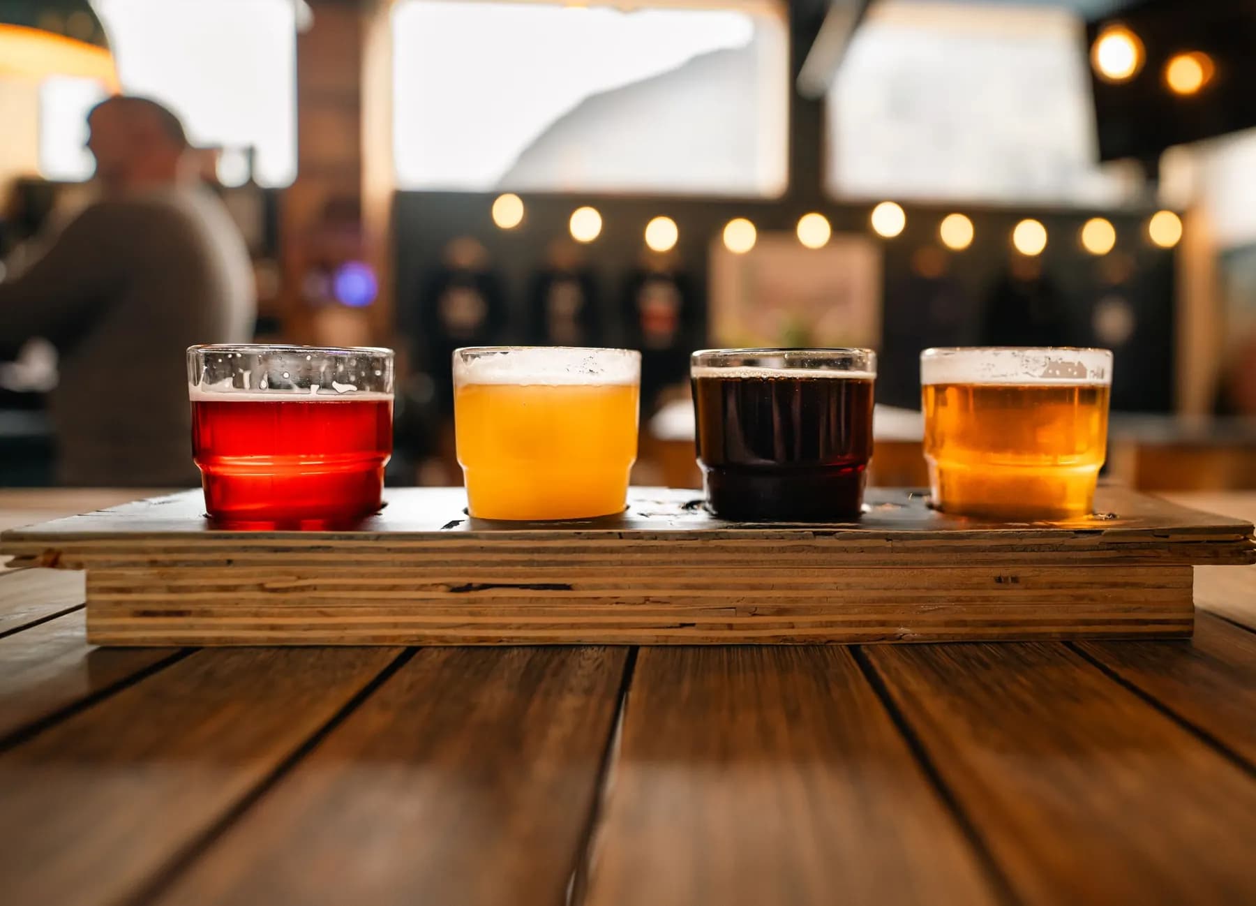 Quatre verres de bière de couleurs différentes, servis sur une planche en bois, sur la table d’un bar aux guirlandes allumées.