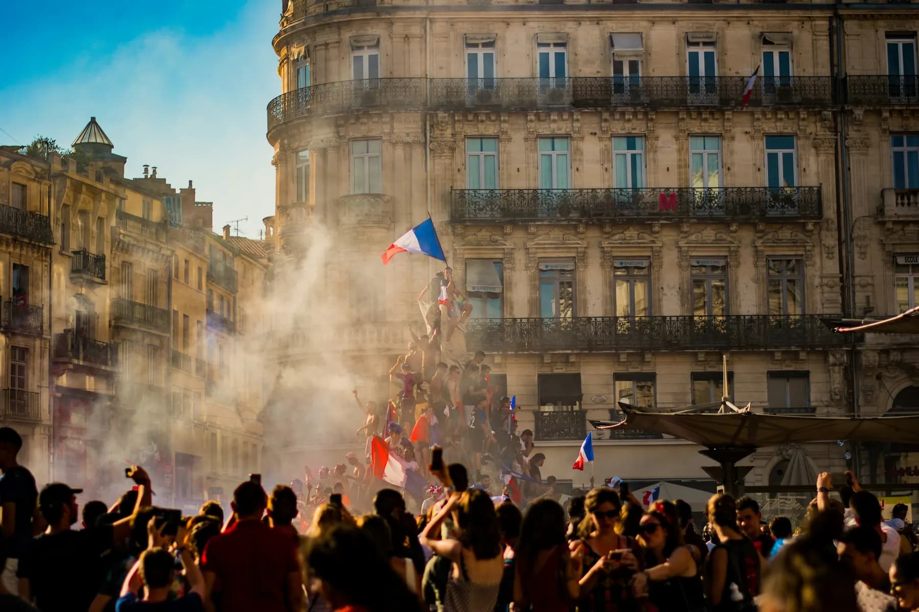 Une place pleine de monde, des drapeaux tricolores et de la fumée, devant de grands immeubles.