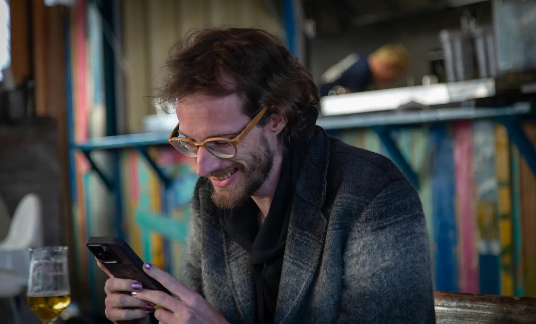 Un homme sourit à son téléphone, attablé dans un bar, une bière posée à côté de lui.