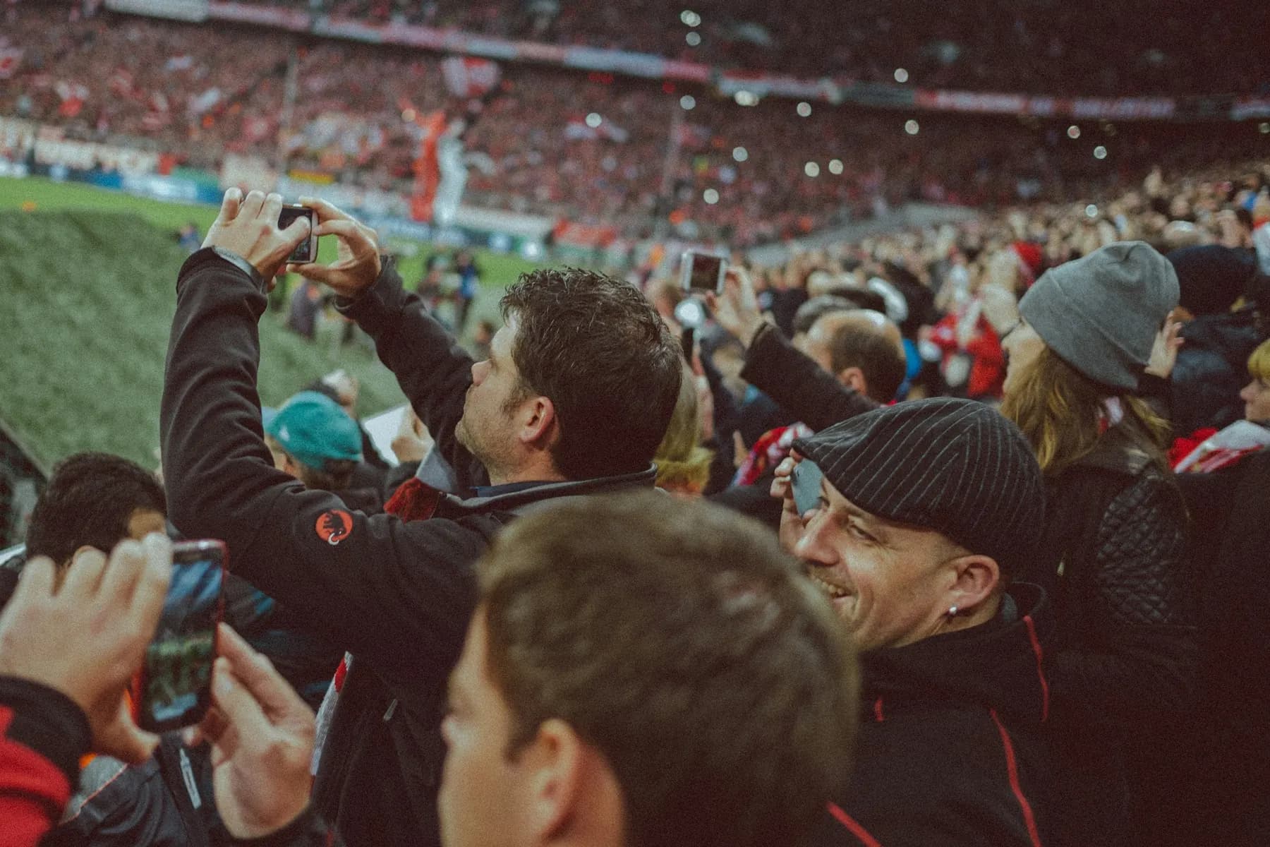 Des supporters filment le match avec leur téléphone, en tribune, en souriant.