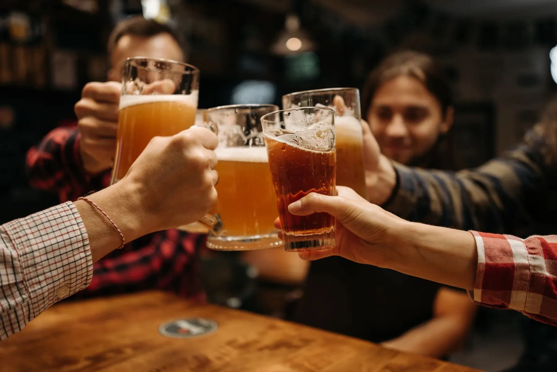 Des amis trinquent, chopes de bière levées, autour d’une table de bar.