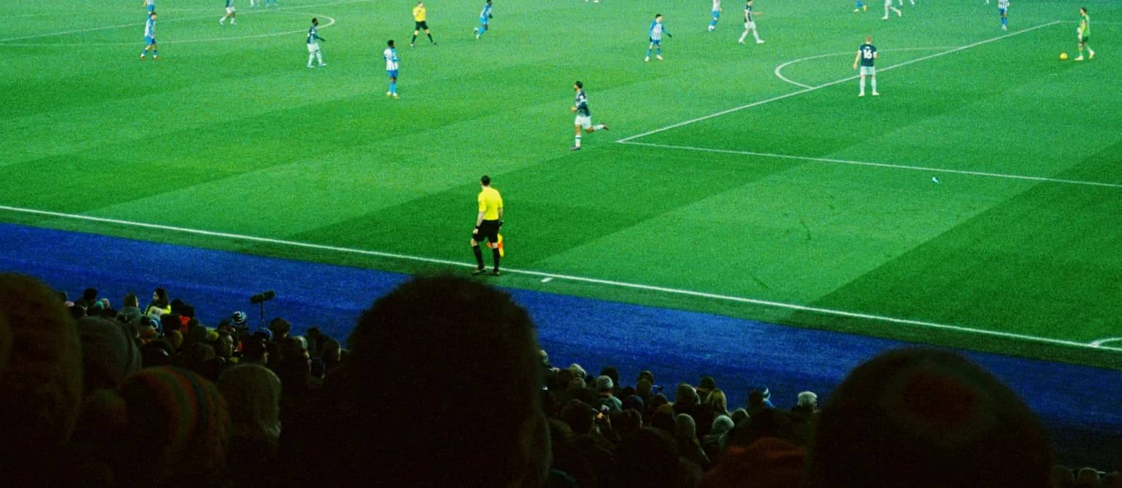 La pelouse d’un stade la nuit, sous les projecteurs, et le public dans l’ombre au premier plan.