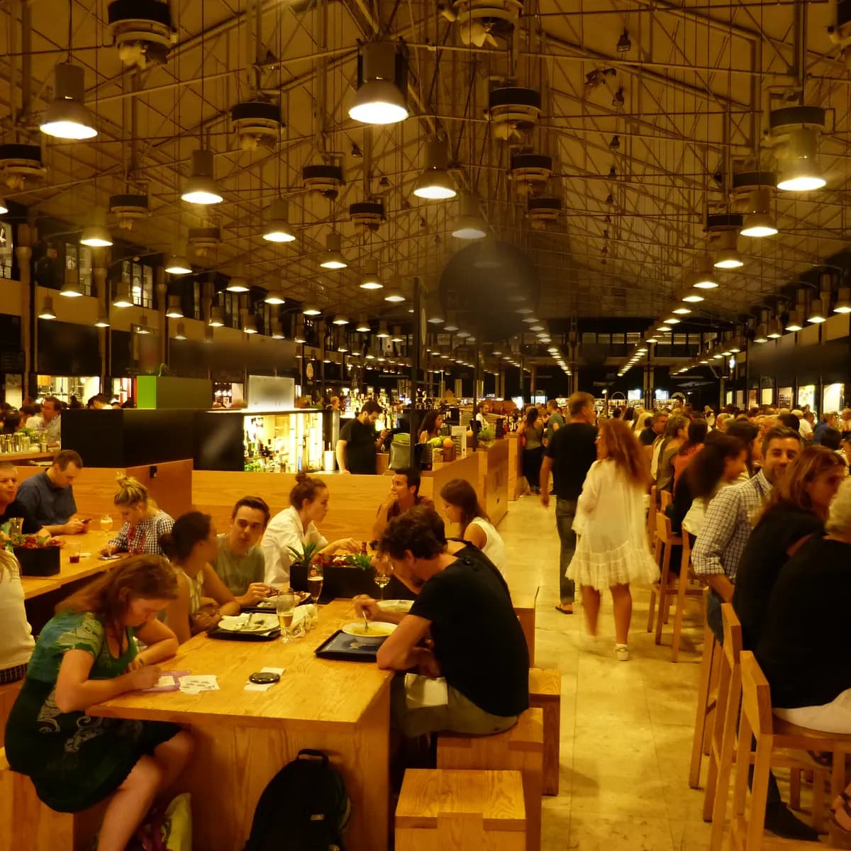 Le soir, une halle gourmande pleine de monde : des convives dînent aux longues tables de bois, entre les stands, sous des dizaines de suspensions allumées et la charpente de fer.
