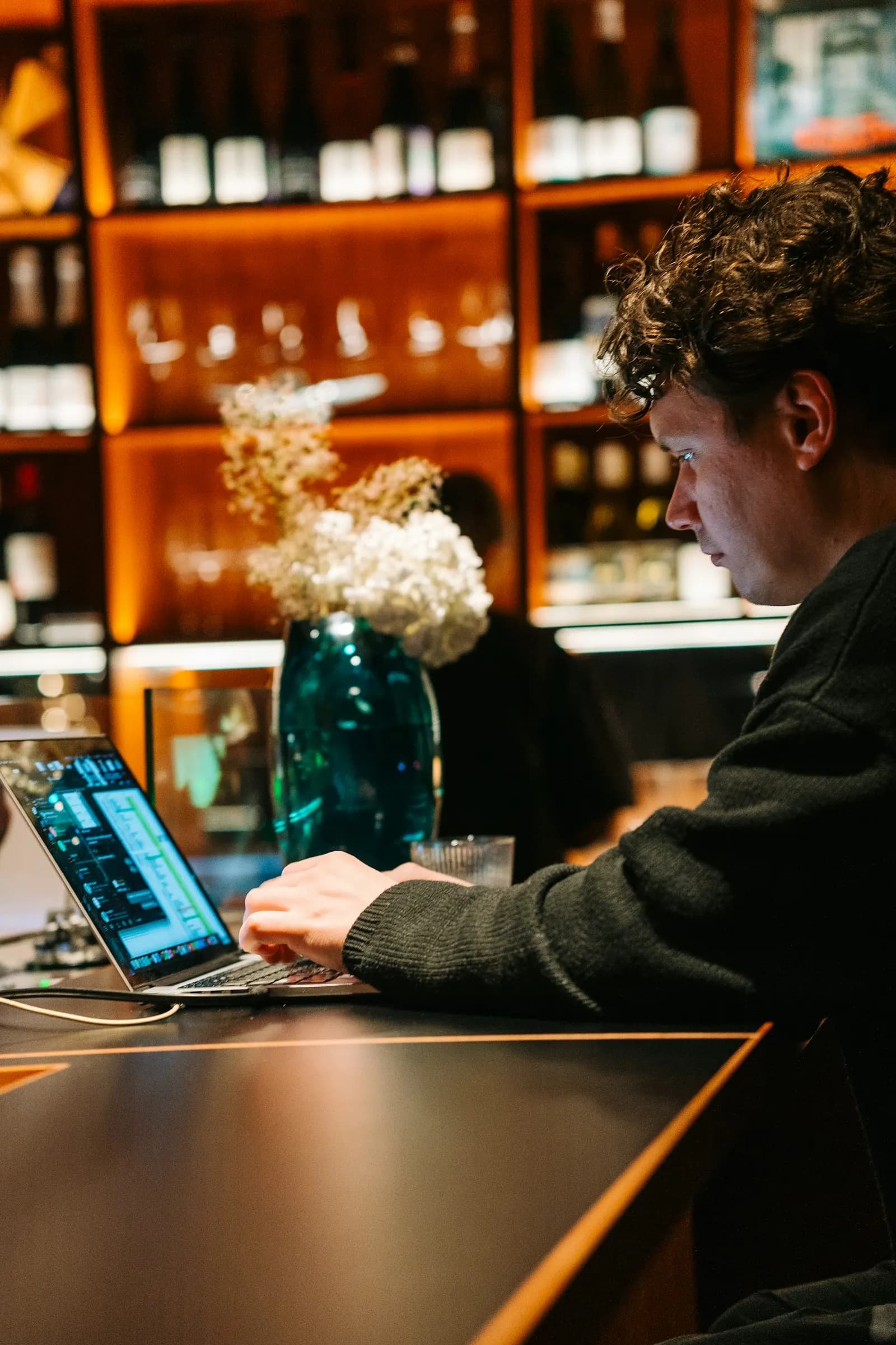 Au comptoir d’un bar, un homme travaille sur son ordinateur portable, devant des étagères de bouteilles éclairées.