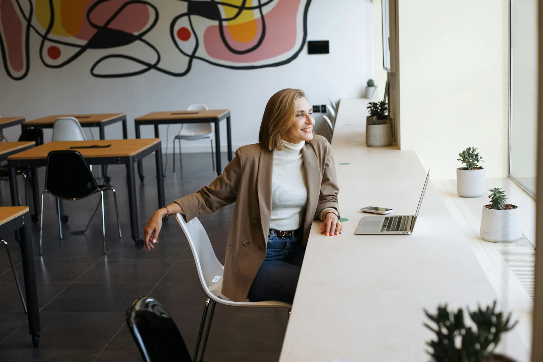 Une femme, son ordinateur portable ouvert devant elle, sourit en regardant par la fenêtre d’une salle claire et vide, en journée.
