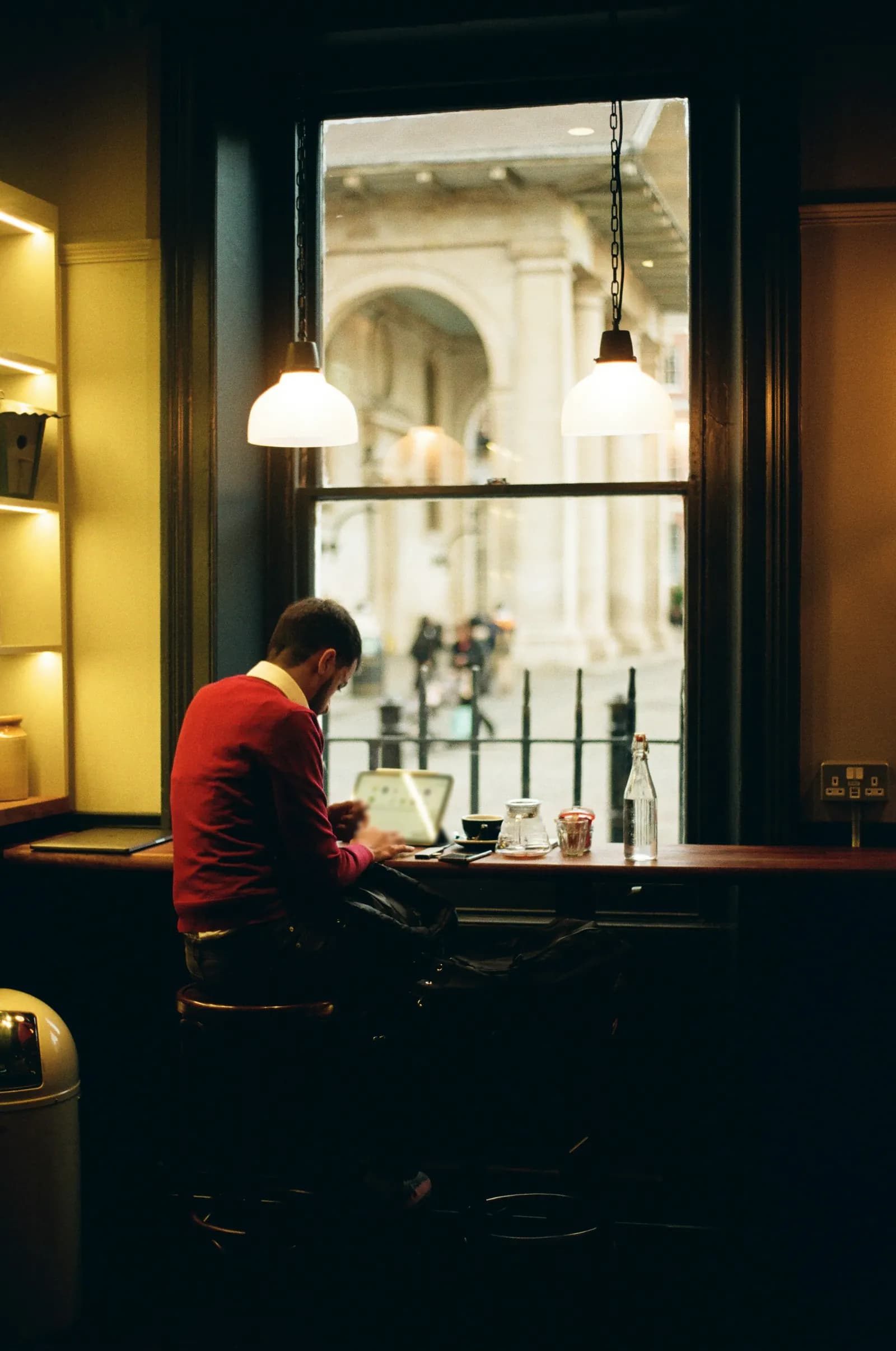 Un homme lit sur une tablette au comptoir sous la fenêtre d’un café, une tasse posée à côté, en fin de journée.