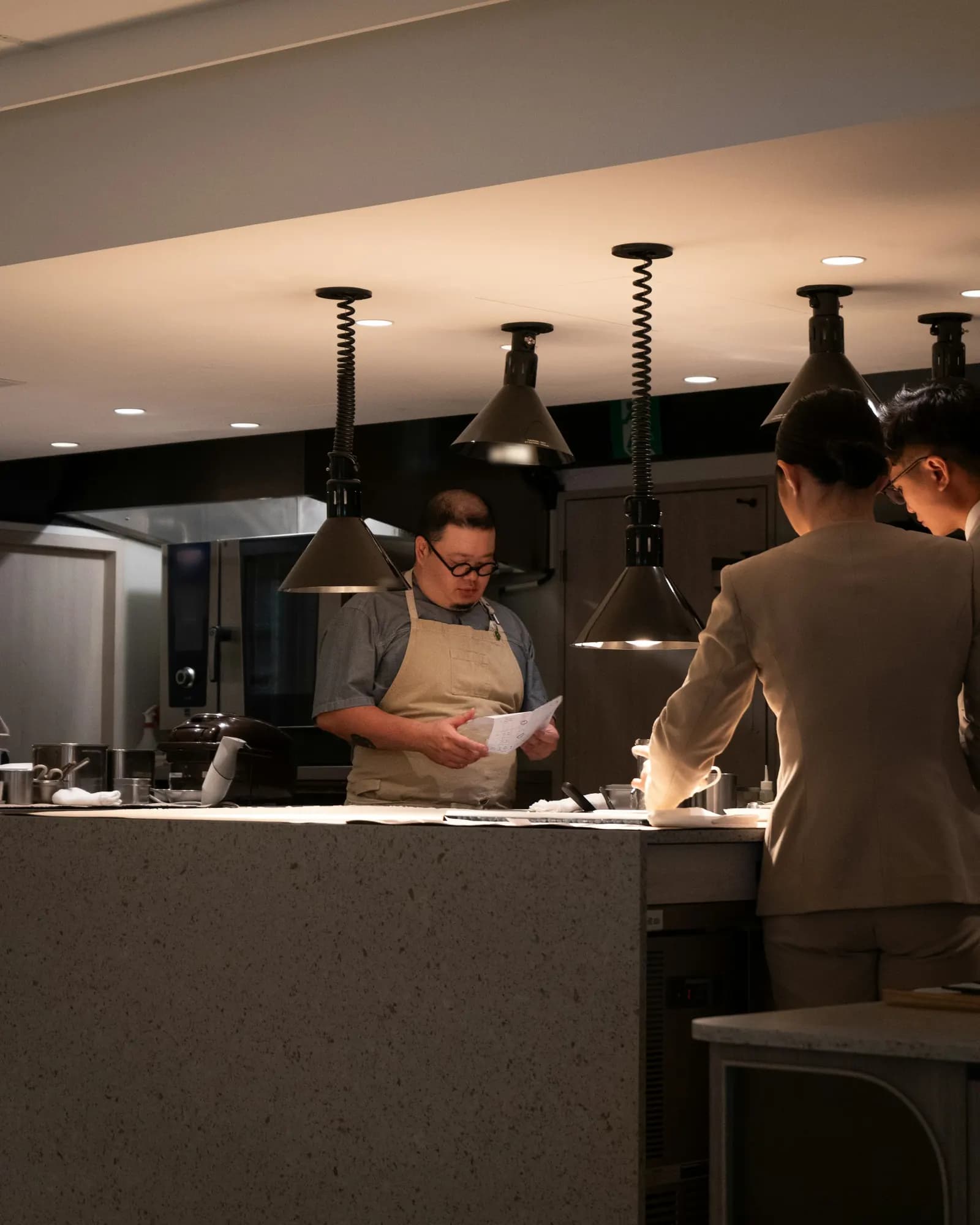 Le chef lit une feuille au passe de sa cuisine ouverte, face à deux membres de la salle, avant le service.