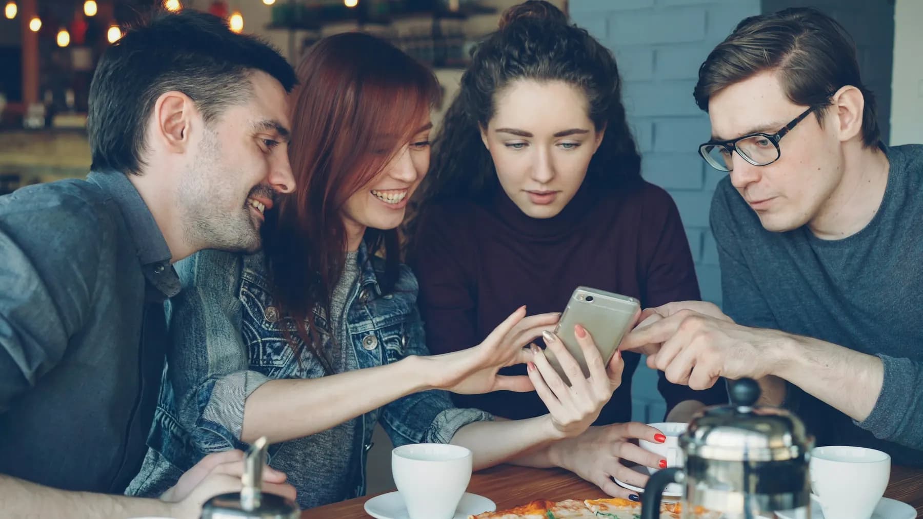 Quatre amis attablés se penchent ensemble sur un téléphone ; deux d’entre eux rient.