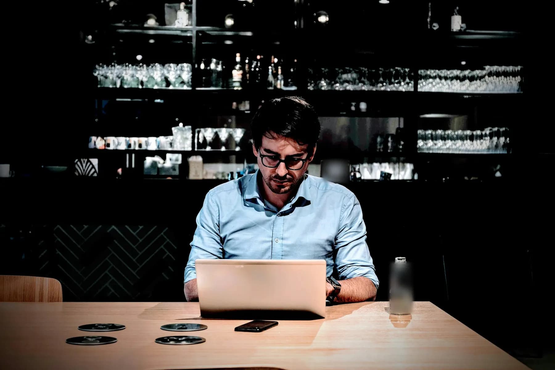 Un homme travaille sur son ordinateur portable à une table de bois, seul dans un bar sombre, devant les étagères de verres et de bouteilles.