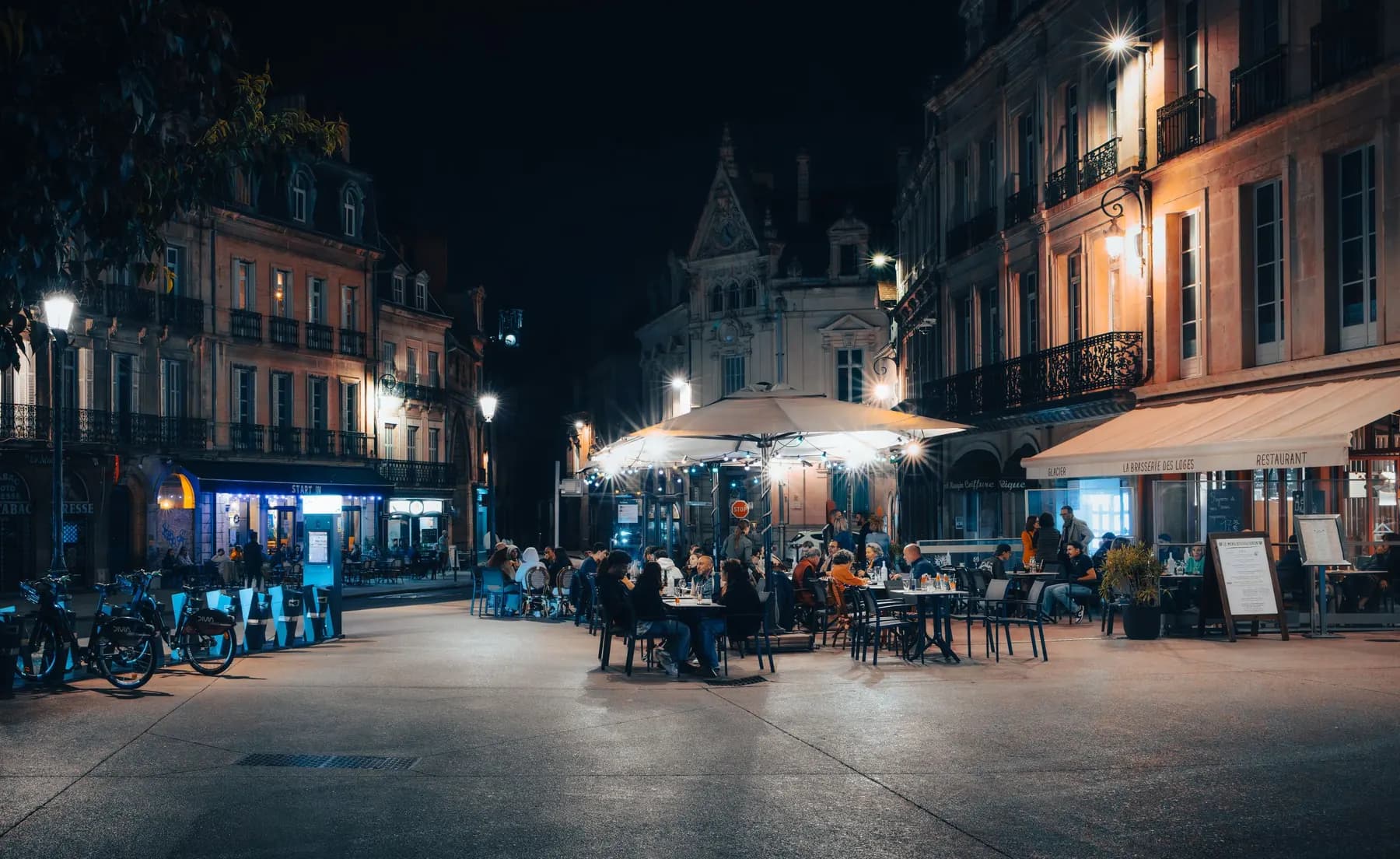 Une place de centre-ville, la nuit : les terrasses d’un bar, d’un café et d’une brasserie sont pleines.