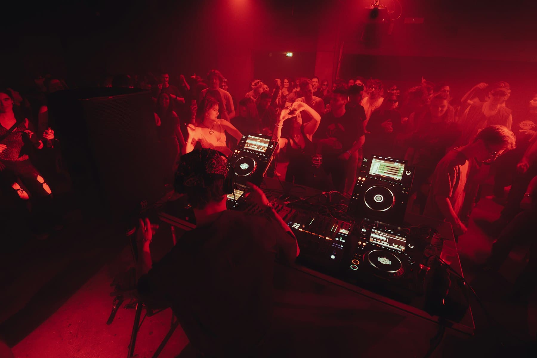 Une piste bondée devant les platines d’un DJ, sous une lumière rouge.