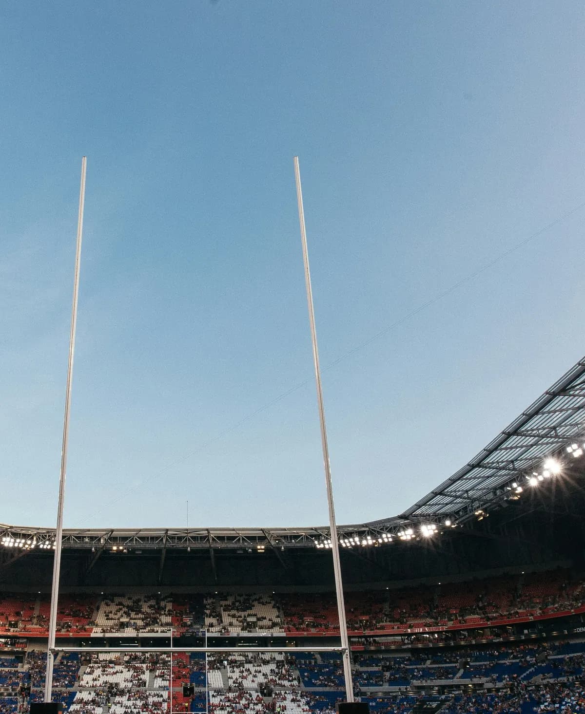 Les poteaux de rugby se dressent devant les tribunes d’un grand stade, sous le toit éclairé.