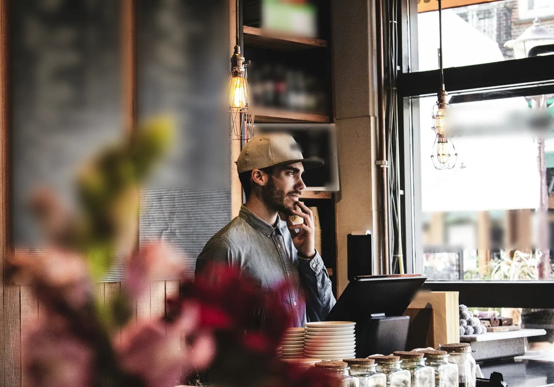 Derrière le comptoir de son café, un homme en casquette et chemise en jean réfléchit, la main au menton, à côté de sa caisse et d’une pile d’assiettes.