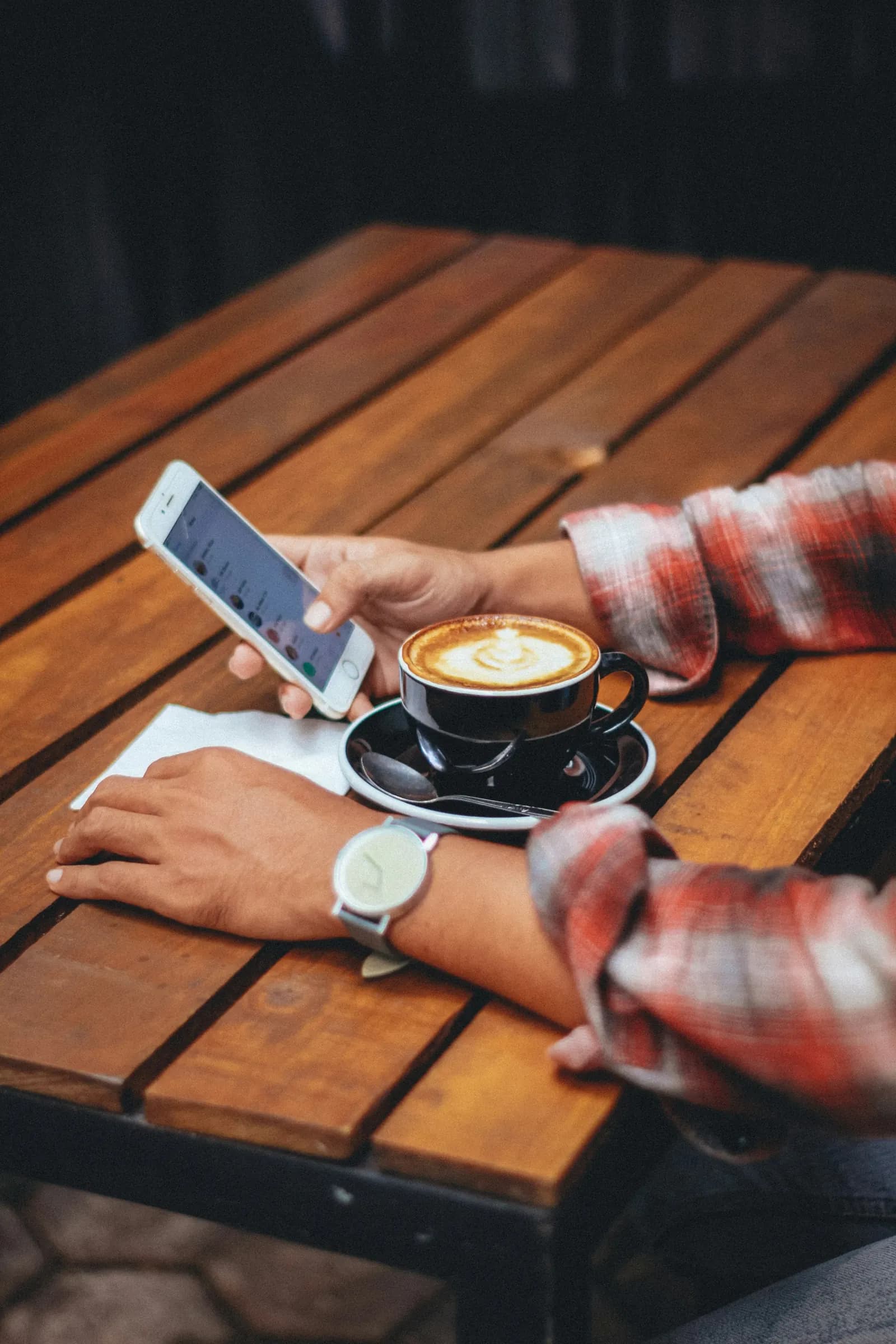 Une main tient un téléphone ouvert sur ses messages, à côté d’un cappuccino, sur une table de café en bois.