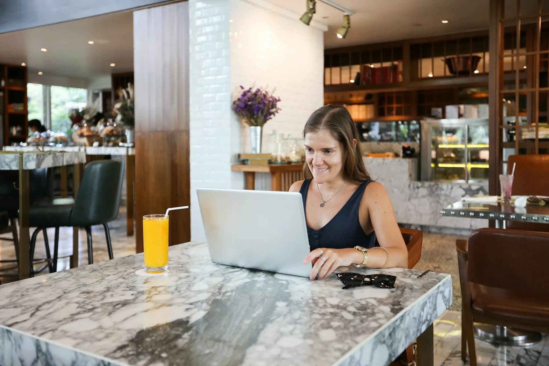 Une femme écrit sur un ordinateur portable à une table en marbre d’un restaurant clair, un jus d’orange à côté.