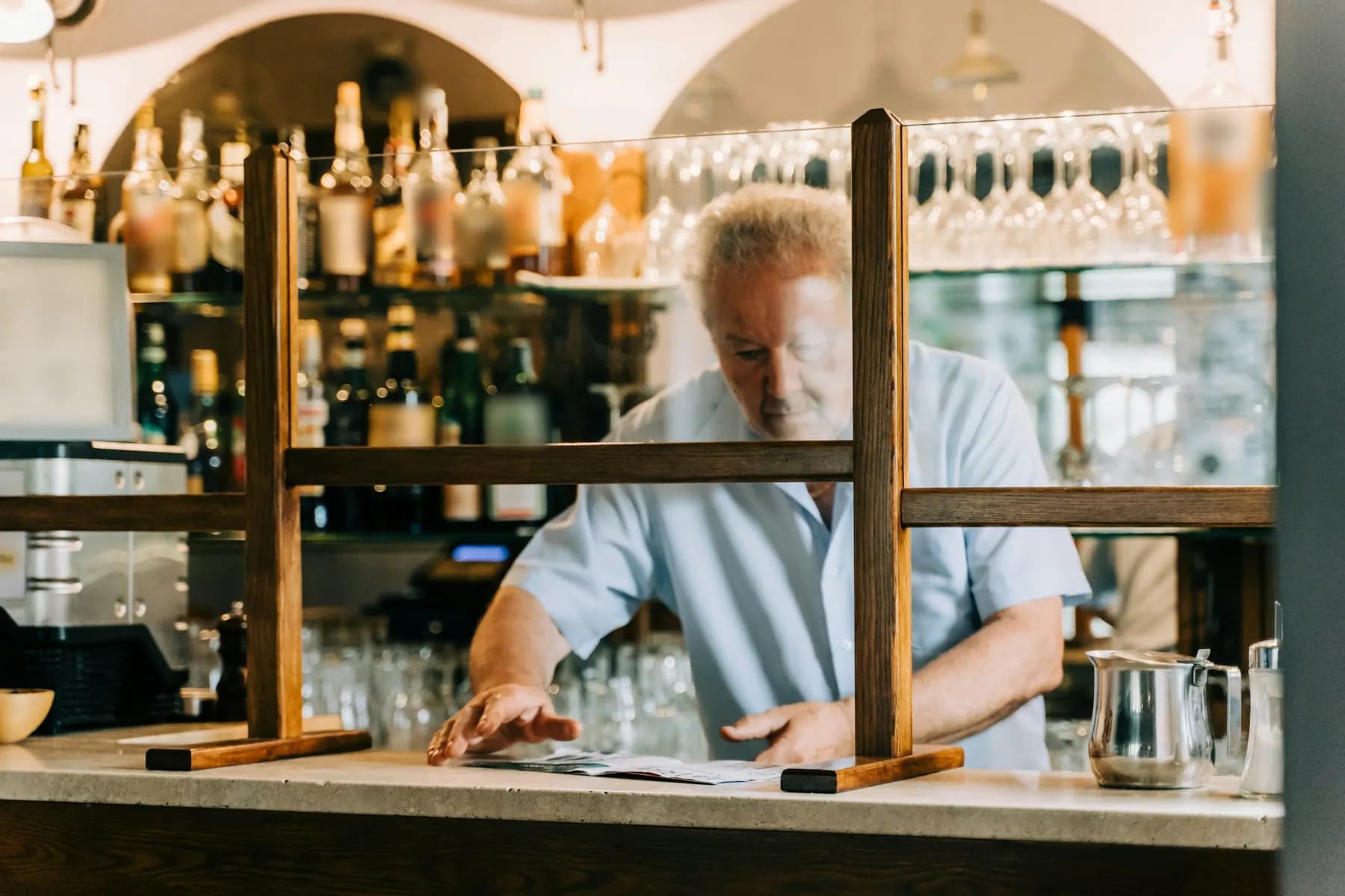 Un patron de bar consulte ses feuilles sur le comptoir, devant les étagères de bouteilles.