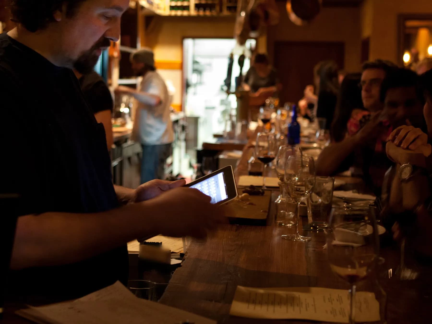 Le soir, derrière le long comptoir de bois d’un bar à charcuterie, un serveur saisit une commande sur la tablette de caisse, face aux clients accoudés.