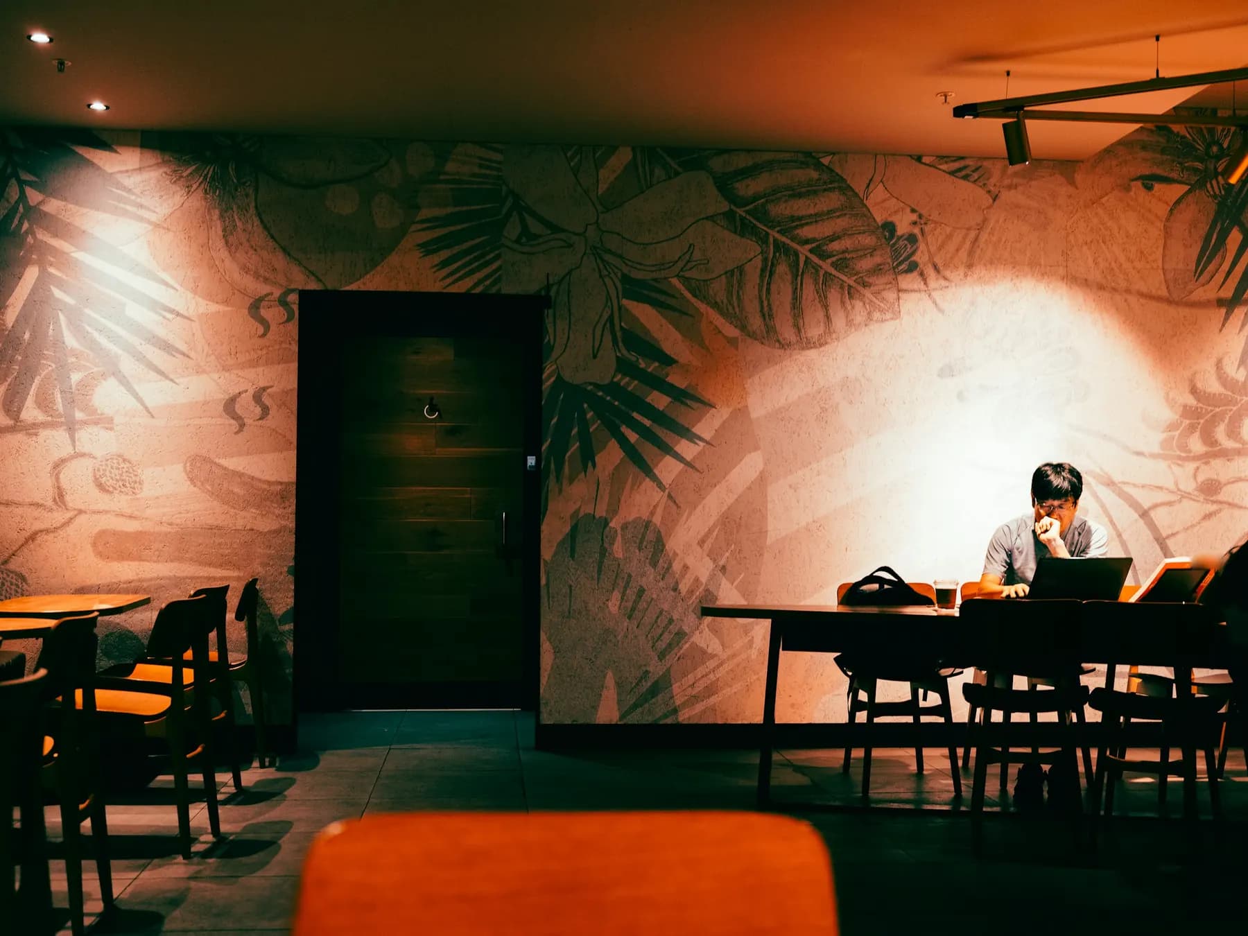 Un homme travaille seul sur un ordinateur portable au fond d’une salle de café vide et tamisée, après la fermeture.
