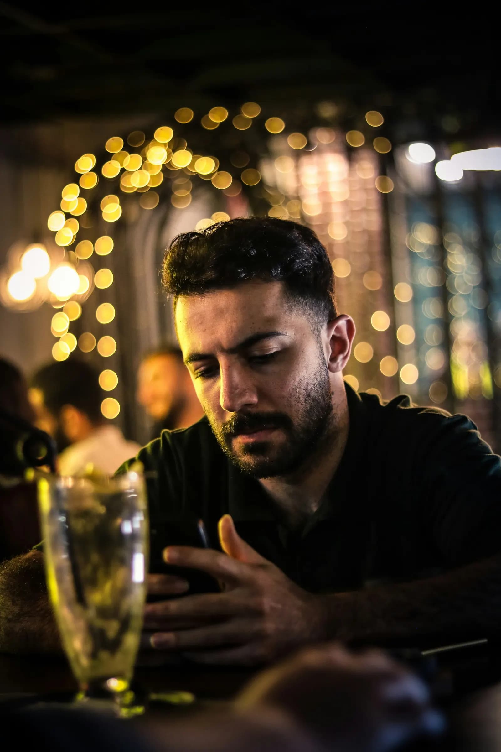 Un homme seul à une table de bar regarde son téléphone, le soir, devant des guirlandes lumineuses.