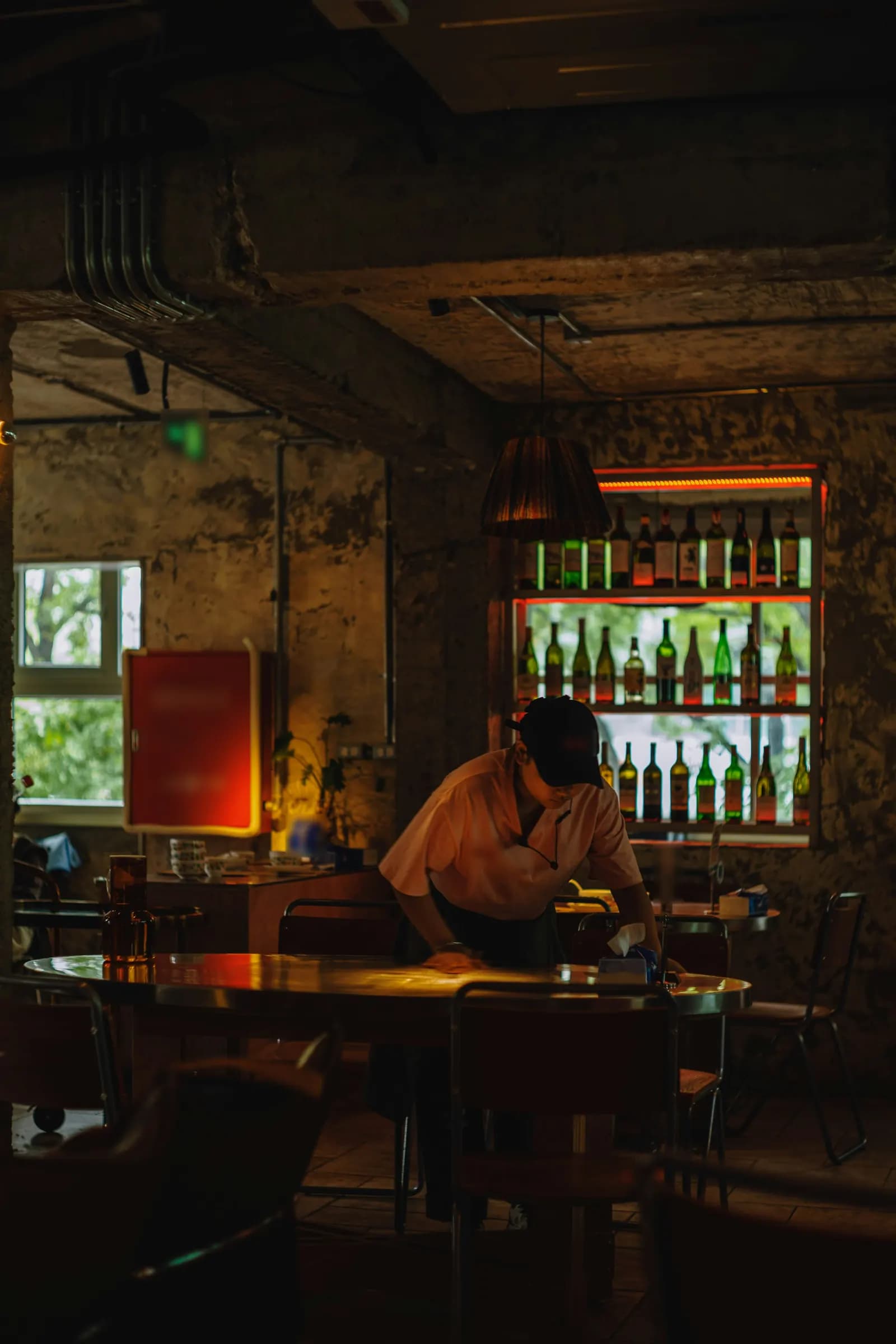 Un employé essuie une table dans un bar à la lumière tamisée, devant une étagère de bouteilles éclairée.