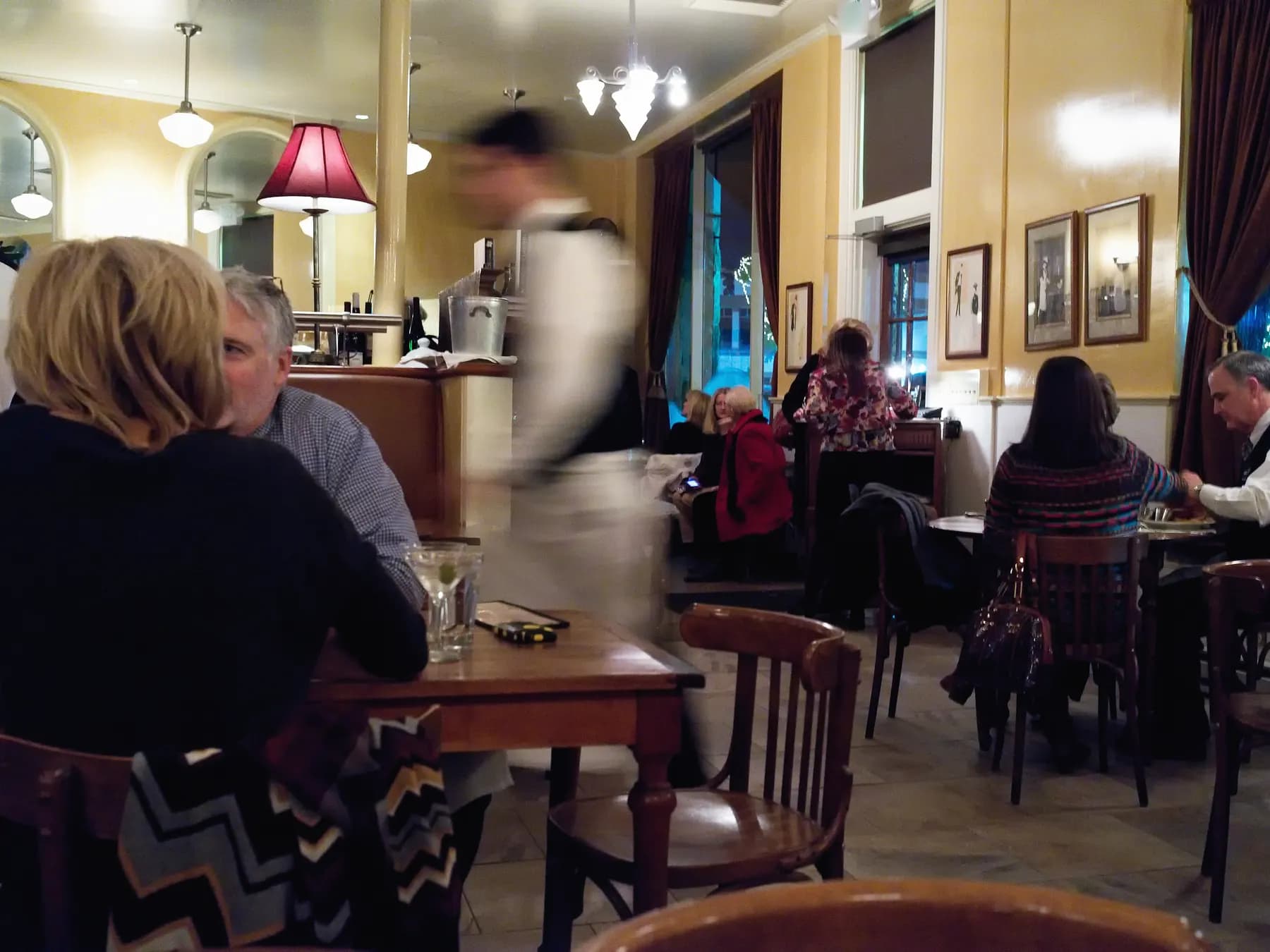 Le soir, dans un bistro aux murs jaunes et aux lampes chaudes, un serveur en tablier traverse la salle entre les tables où dînent les clients.
